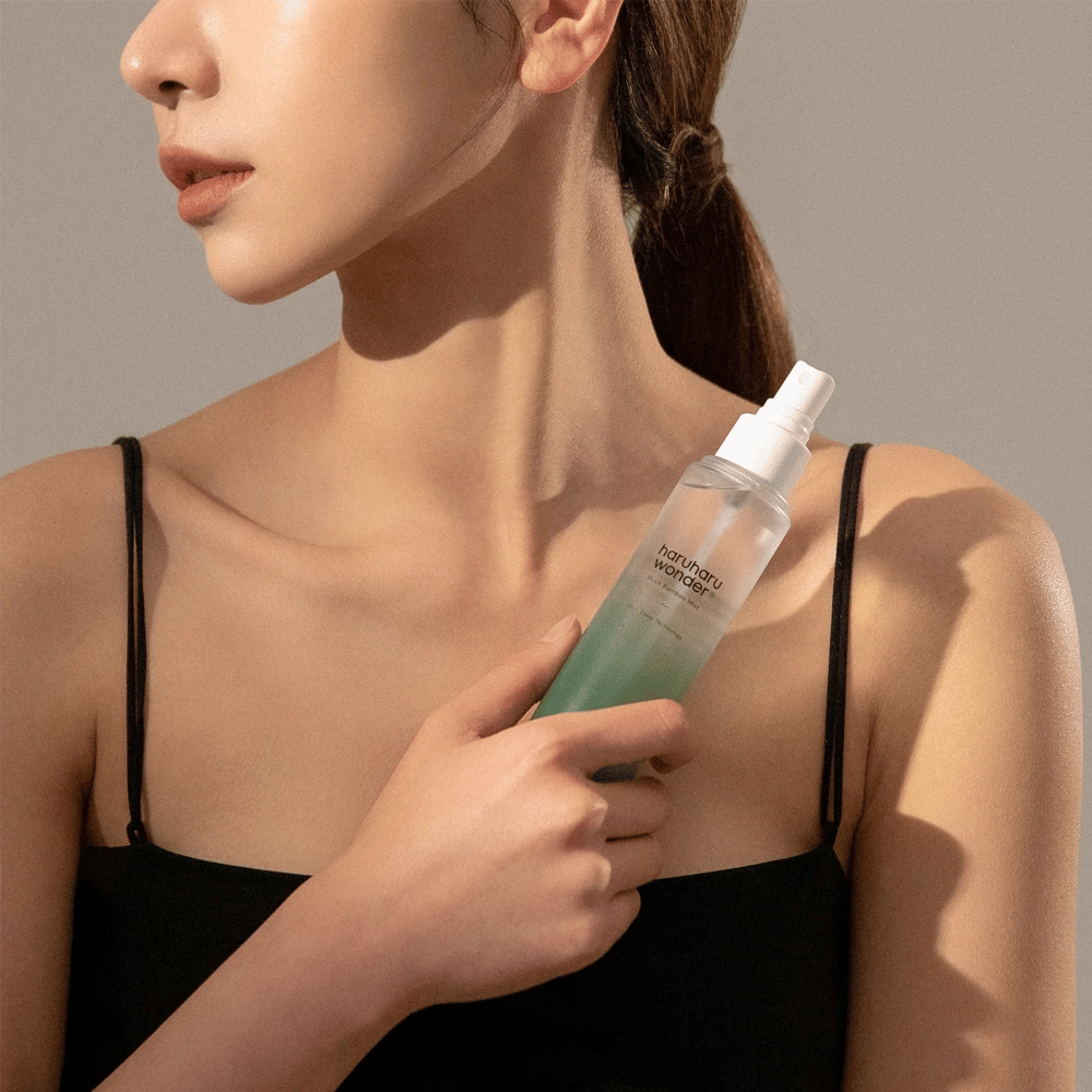 Imagen de HaruHaru Wonder Black Bamboo Mist sostenida por una mujer. El producto está en una botella de spray transparente con un tono verde y una bomba blanca. La mujer tiene un aspecto relajado, lleva una blusa negra y se enfoca en usar la bruma para dar humedad y brillo a la piel. La botella es parte de una rutina de cuidado de la piel.