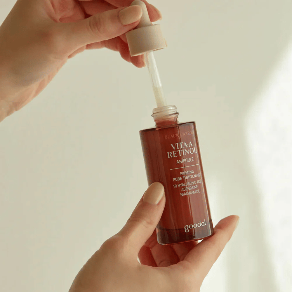 Una mano sostiene una botella con Goodal Black Carrot Vita-A Retinol Firming Ampoule. La etiqueta de la botella muestra el nombre y las propiedades del producto, incluyendo "Firming Pore Tightening" e ingredientes como 10% ácido hialurónico, adenosina y niacinamida. El gotero es de color beige.