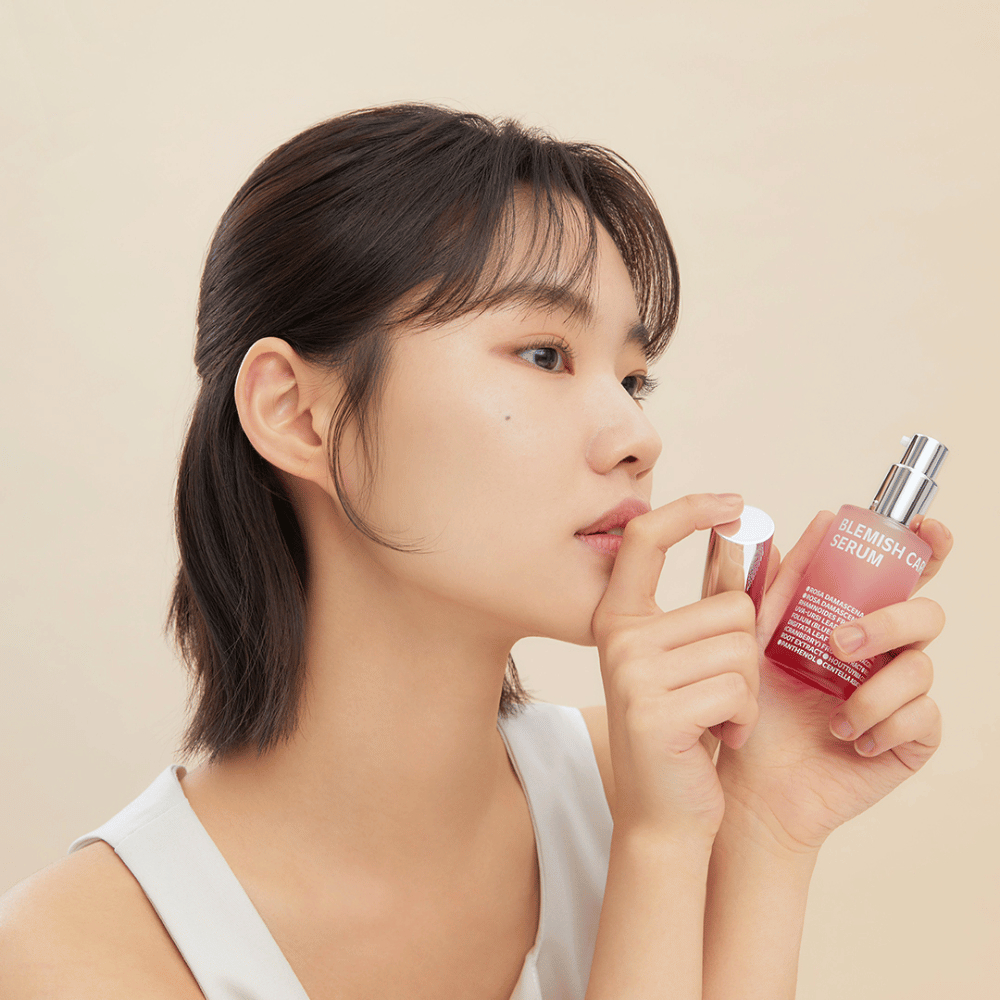 Una mujer con cabello oscuro sostiene una botella de Blemish Care Serum en una mano y quita el tapón con la otra. Tiene una expresión facial neutral y el fondo es de un beige claro, lo que da una impresión suave y minimalista.
