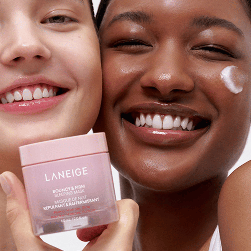 Dos personas sonríen y sostienen Laneige Bouncy & Firm Sleeping Mask. Una de ellas ha aplicado la mascarilla en su mejilla, y el producto se ve claramente en sus manos. La mascarilla está en un tarro rosa y se comercializa como "Volumen, Hidratación, Brillo."