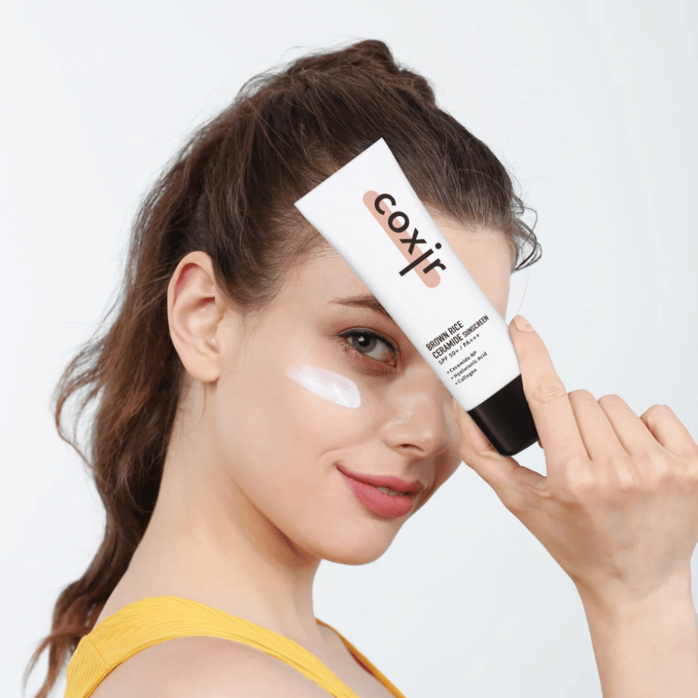 Mujer con crema solar aplicada en la mejilla sostiene un tubo Coxir Brown Rice Ceramide Sunscreen, SPF 50+ PA+++, hacia la frente y sonríe ligeramente.
