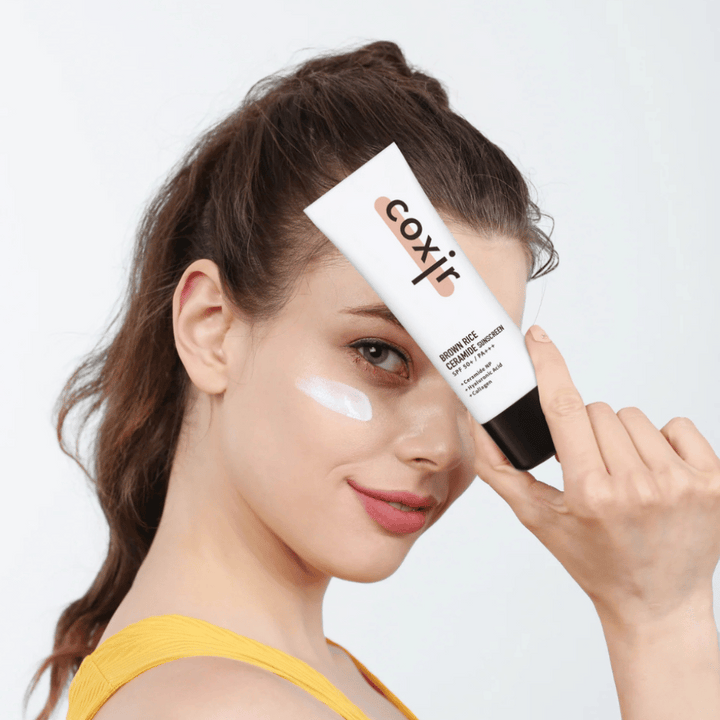 Kvinna med solkräm applicerad på kinden håller upp en tub Coxir Brown Rice Ceramide Sunscreen, SPF 50+ PA+++, mot pannan och ler lätt.