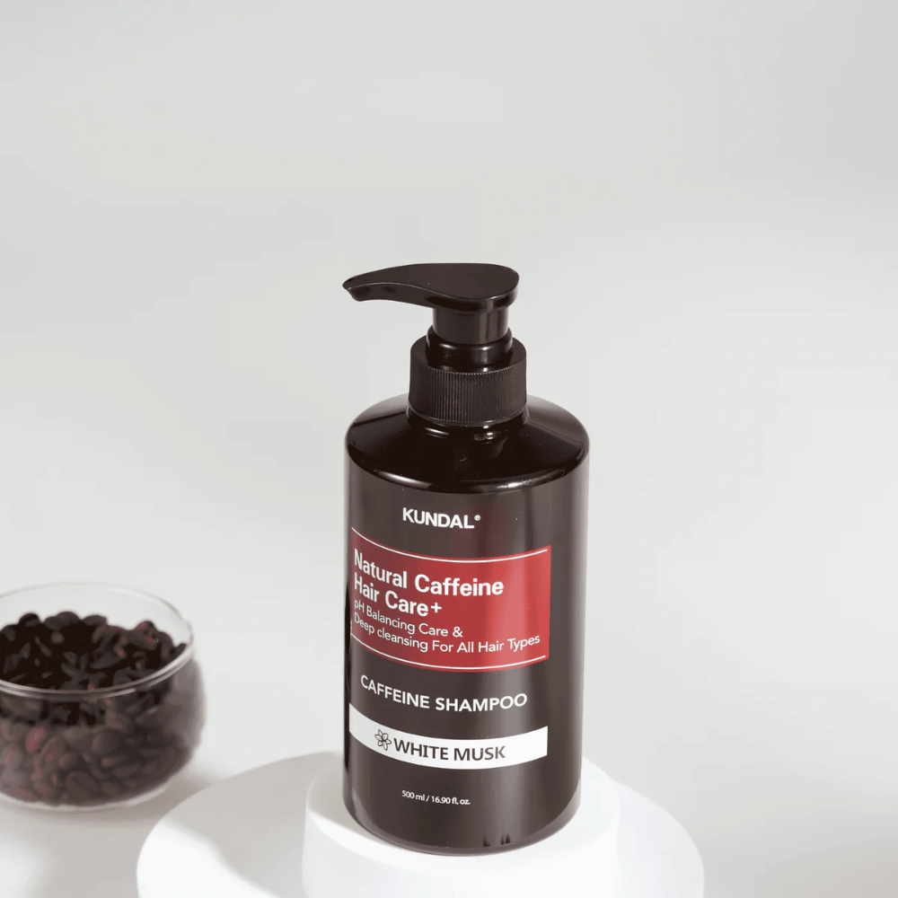 KUNDAL Natural Caffeine Hair Care+ Shampoo White Musk en botella negra con bomba, 500 ml, colocada junto a un cuenco con granos de café.