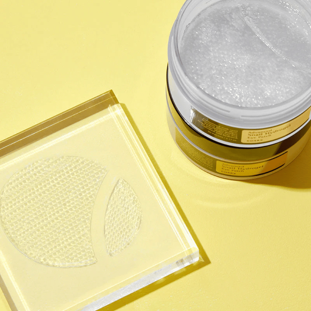 Un tarro abierto COSRX Advanced Snail Hydrogel Eye Patch está sobre un fondo amarillo junto a un posavasos transparente donde se colocan dos parches para ojos.