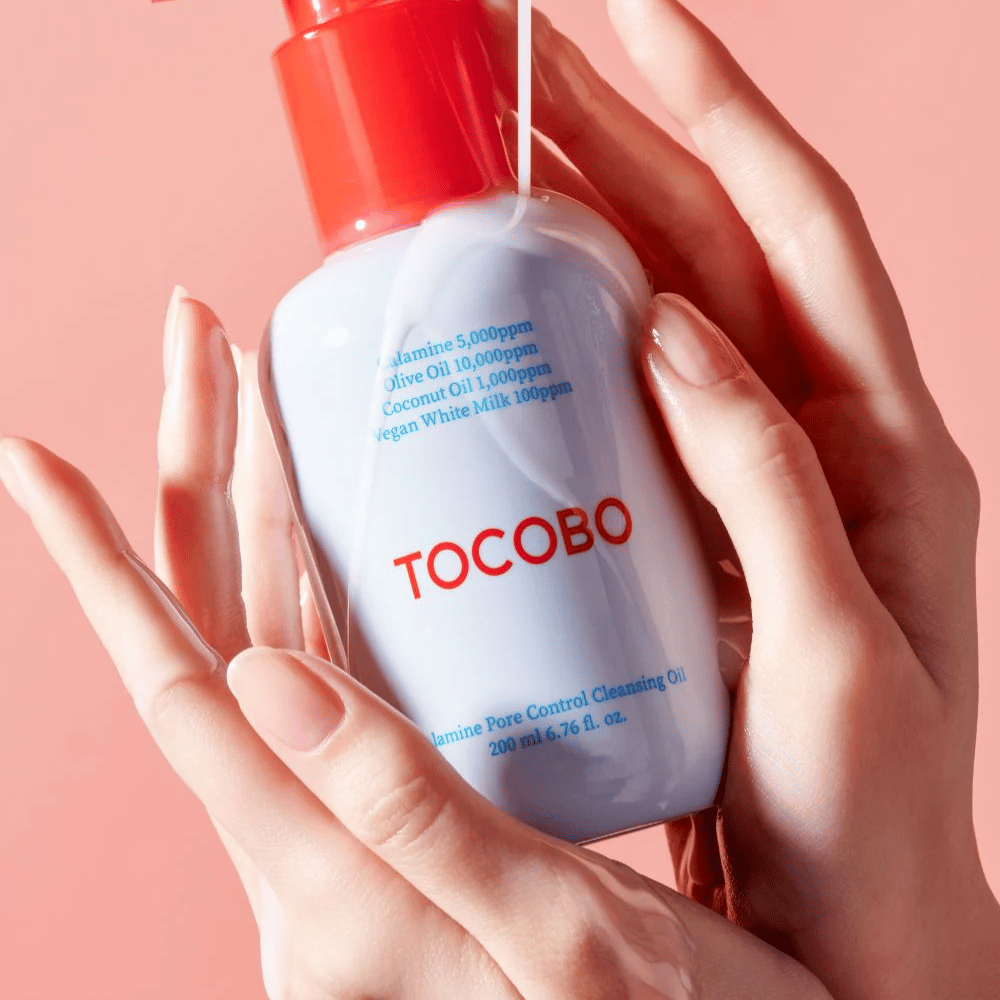 TOCOBO Calamine Pore Control Cleansing Oil sostenido en manos contra un fondo rosa, la bomba roja de la botella y la lista de ingredientes se ven claramente.