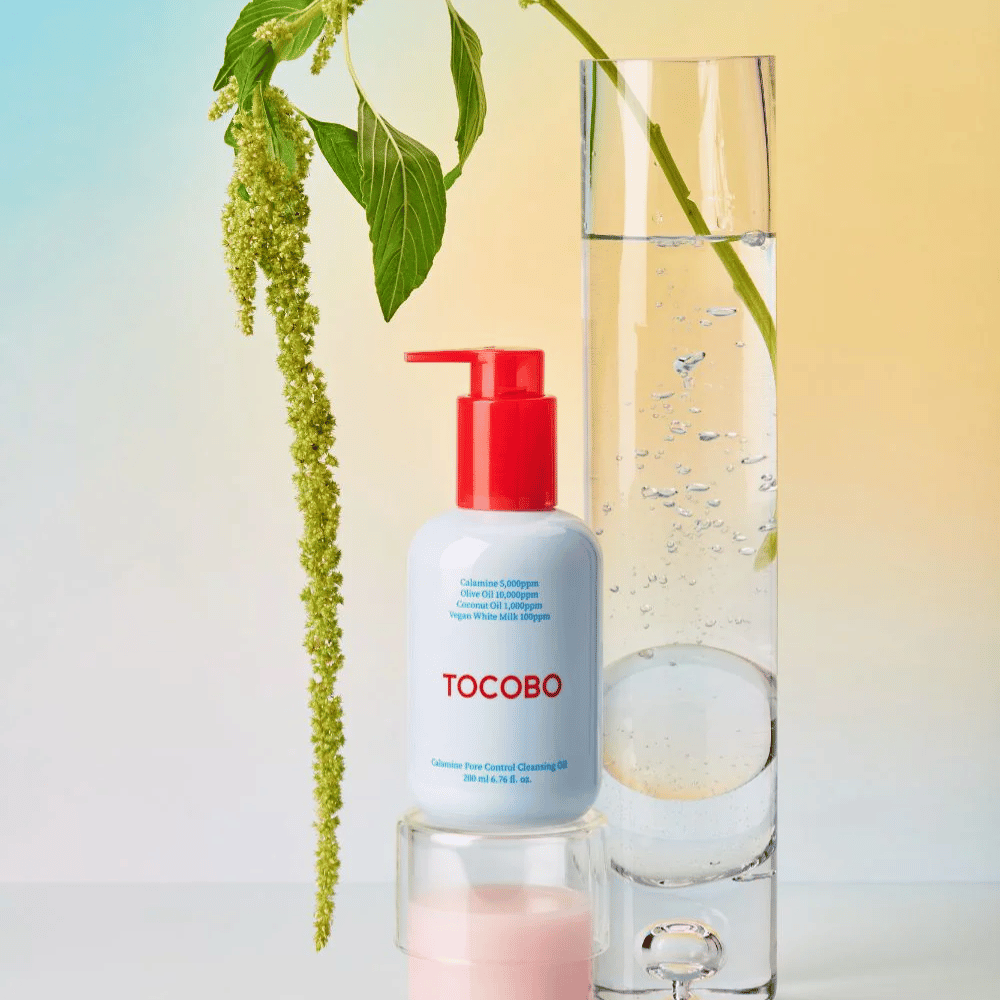 TOCOBO Calamine Pore Control Cleansing Oil con bomba roja está al lado de un tubo de vidrio alto lleno de agua y una planta verde, contra un fondo de color pastel claro.