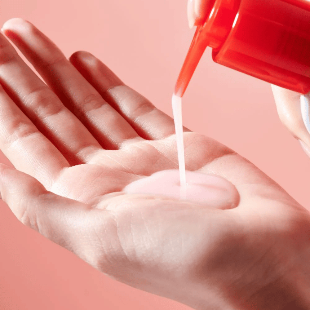Se aplica un aceite limpiador rosa claro desde una botella de bomba roja en una mano abierta contra un fondo rosa.