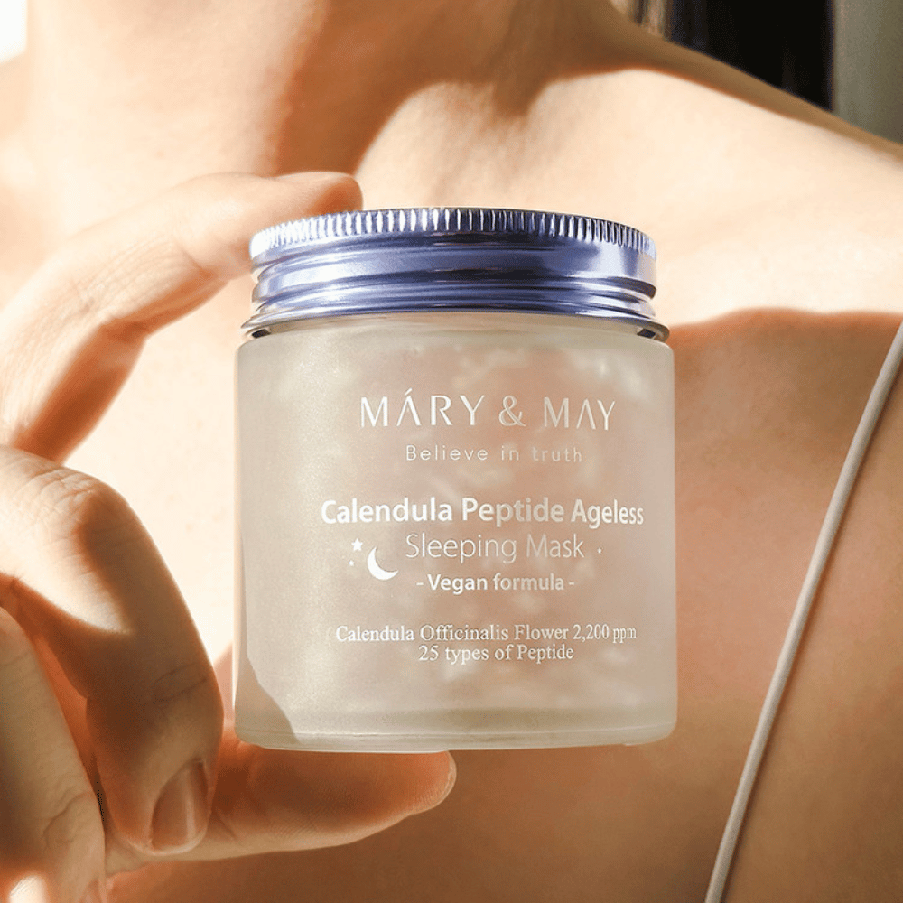 Una mujer sostiene un frasco de MARY & MAY Calendula Peptide Ageless Sleeping Mask. El frasco es transparente con una tapa lila y el texto "Calendula Peptide Ageless Sleeping Mask" se ve claramente. El producto es una mascarilla vegana para dormir con caléndula y péptidos, destinada a darle a la piel un brillo juvenil durante la noche.
