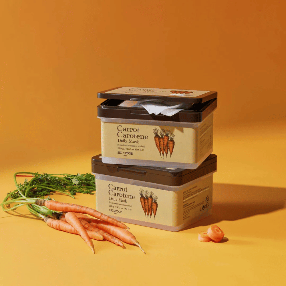La imagen muestra tres paquetes apilados de "Carrot Carotene Daily Mask" de SKINFOOD, colocados sobre un fondo naranja junto con zanahorias frescas. Cada caja contiene mascarillas faciales enriquecidas con β-caroteno de aceite de semilla de zanahoria, diseñadas para uso diario para nutrir y cuidar la piel.