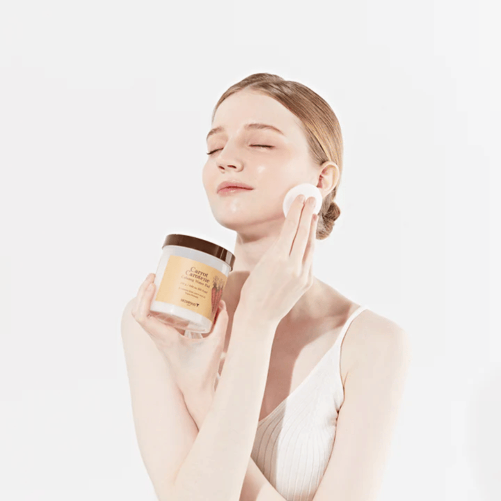Una mujer con el cabello recogido aplica un producto de cuidado facial en su rostro con una almohadilla de algodón. Ella sostiene un gran frasco etiquetado "Carrot Carotene Calming Water Pad". Sus ojos cerrados y su expresión pacífica transmiten una sensación de relajación y disfrute de la rutina de cuidado facial.