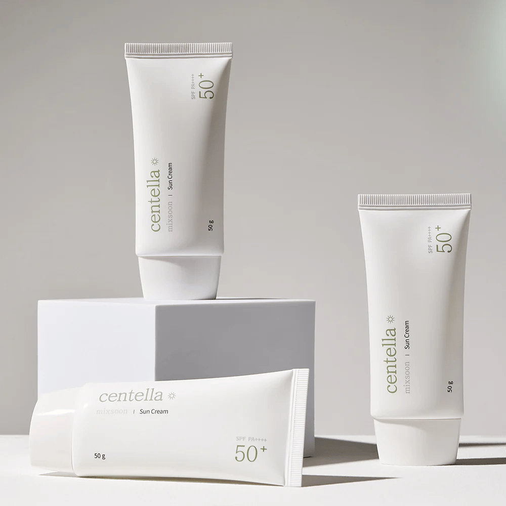 Tres paquetes de Centella Sun Cream SPF 50+ PA++++ dispuestos estéticamente en podios blancos. Dos tubos están de pie y uno está acostado, todos con un diseño blanco, minimalista y detalles de texto en verde.