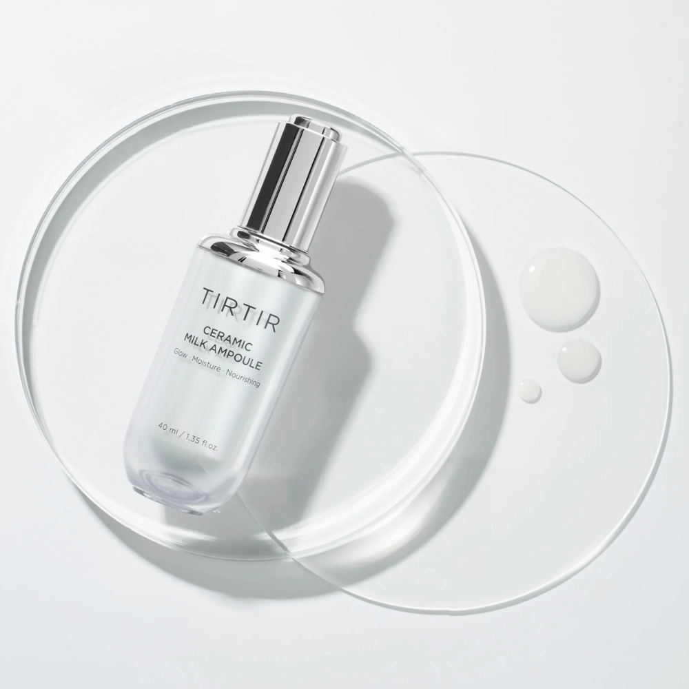 Una botella transparente TIRTIR Ceramic Milk Ampoule sobre una placa de vidrio con gotas, contra un fondo blanco, destaca brillo, humedad y nutrición.