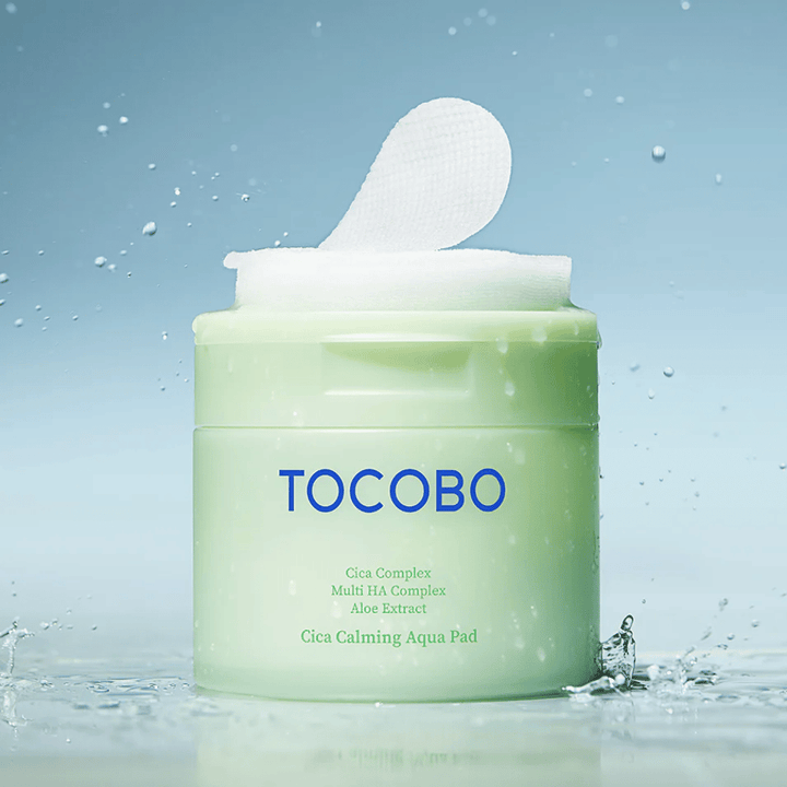Tarro abierto de TOCOBO Cica Calming Aqua Pad donde una almohadilla de tónico sobresale del envase. Las gotas de agua alrededor del producto refuerzan la sensación de hidratación intensa y frescura.