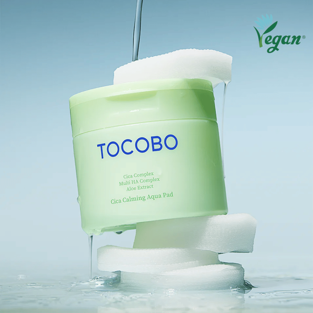 TOCOBO Cica Calming Aqua Pad colocada entre almohadillas de tónico con esencia escurriendo. El símbolo vegano enfatiza la formulación vegana del producto y su perfil respetuoso con la piel.