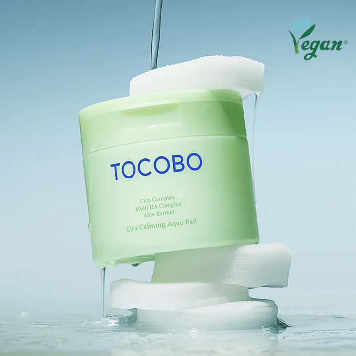 TOCOBO Cica Calming Aqua Pad colocada entre almohadillas de tónico con esencia escurriendo. El símbolo vegano enfatiza la formulación vegana del producto y su perfil respetuoso con la piel.