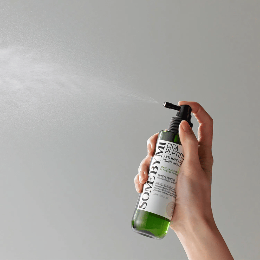 Mano rociando SOME BY MI Cica Peptide Anti Hair Loss Derma Scalp Tonic desde una botella verde con boquilla negra y etiqueta blanca sobre fondo gris.