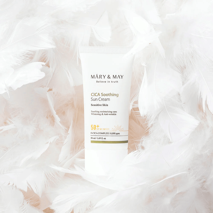 Mary&May CICA Soothing Sun Cream rodeada de suaves plumas blancas. La imagen transmite una sensación de suavidad, comodidad y una fórmula ligera adaptada para piel sensible.