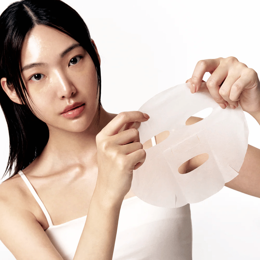 Mujer con piel limpia e hidratada sostiene una Sheet mask blanca frente a ella, lista para aplicarla en el rostro.
