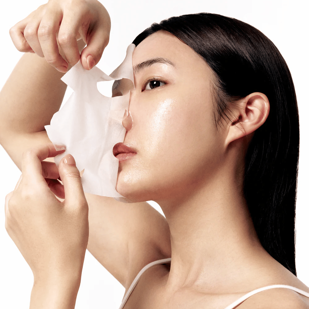 Mujer con piel lisa e hidratada se quita una Sheet mask blanca del rostro, revelando un tono de piel radiante y uniforme.