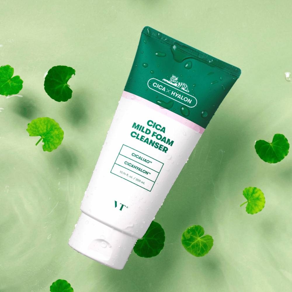 VT Cica Mild Foam Cleanser acostado sobre una superficie verde claro con gotas de agua en el envase y rodeado de hojas verdes de centella asiática, lo que resalta las propiedades calmantes del producto.