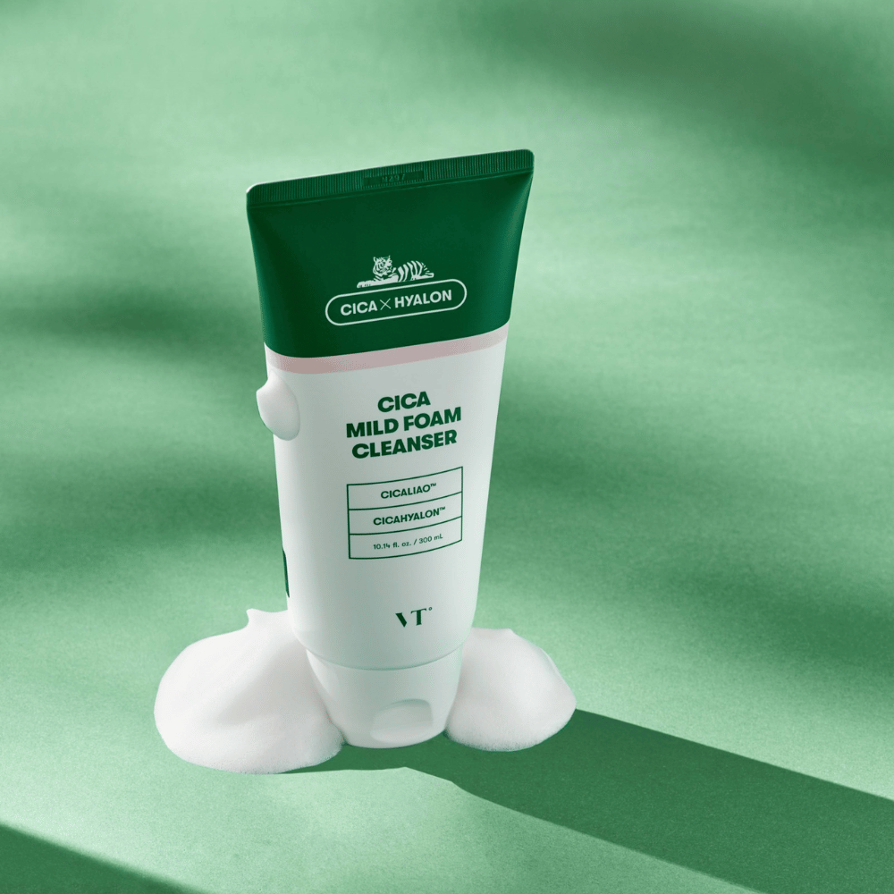 Tubo con VT Cica Mild Foam Cleanser de pie sobre una superficie verde con espuma alrededor de la base. El producto tiene un diseño verde y blanco y se comercializa con CICALIAO™ y CICAHYALON™.