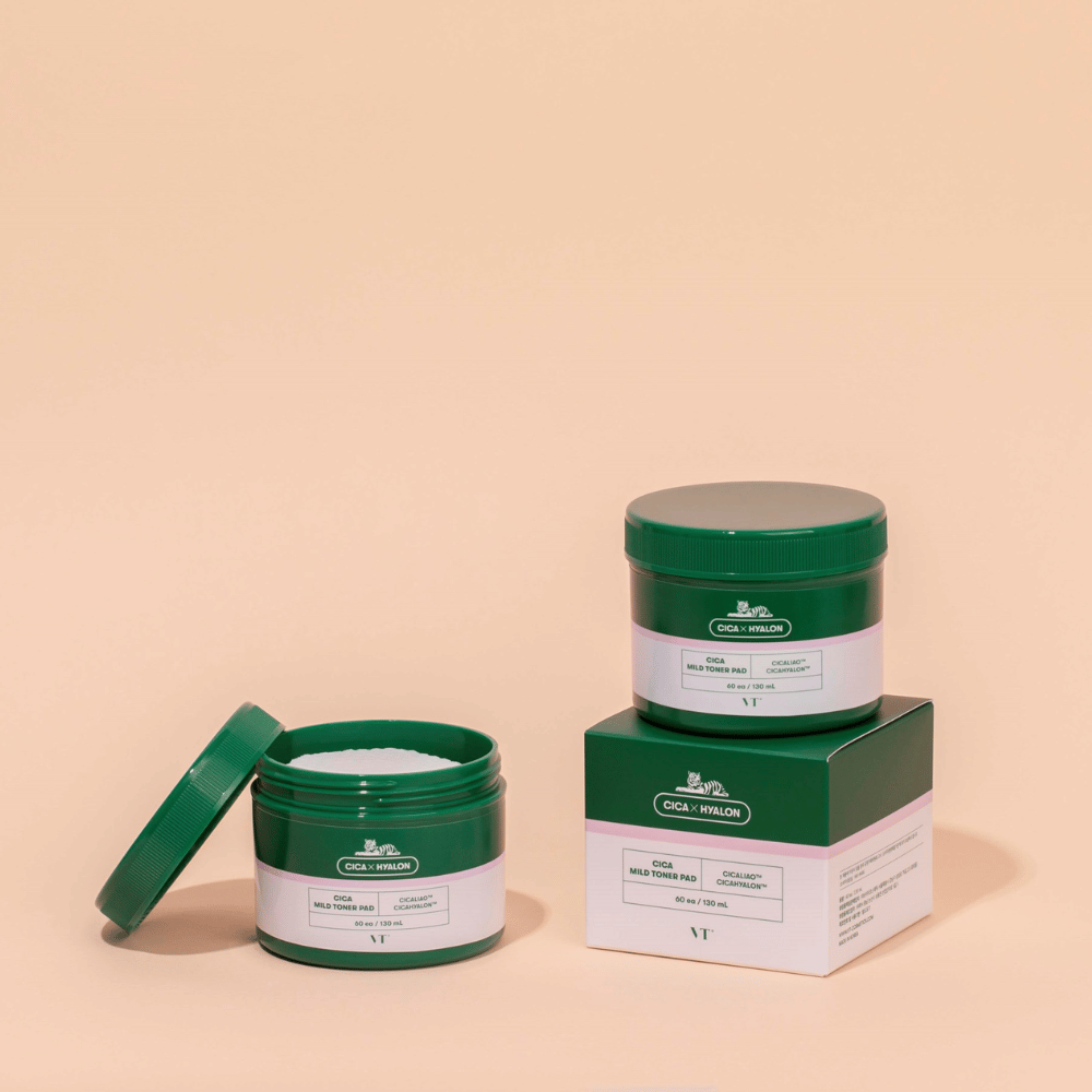 Dos botes verdes con VT Cica Mild Toner Pad, uno abierto que muestra las almohadillas de tónico blancas y uno cerrado colocado sobre su empaque de producto, sobre fondo beige.