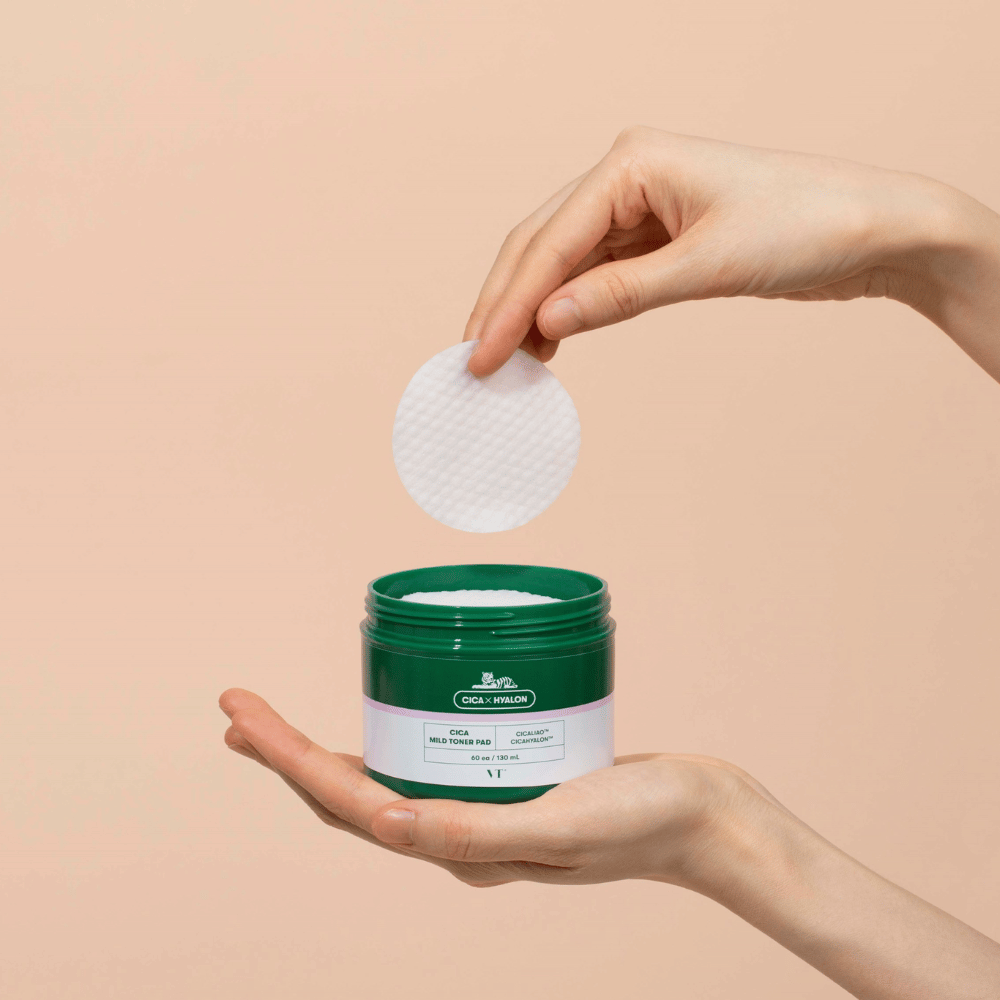 Mano sosteniendo un bote verde con VT Cica Mild Toner Pad y levantando una almohadilla de tónico redonda y blanca sobre el bote contra un fondo beige.