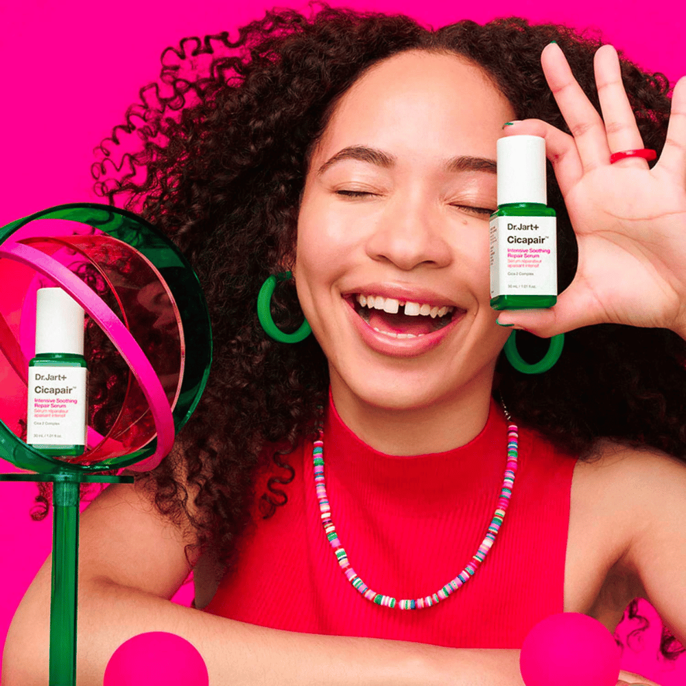 Una mujer sonríe ampliamente mientras sostiene Dr.Jart+ Cicapair Intensive Soothing Repair Serum frente a su rostro. Lleva pendientes verdes y un collar rojo. El fondo es de color rosa claro y está decorado con círculos verdes y rosas.