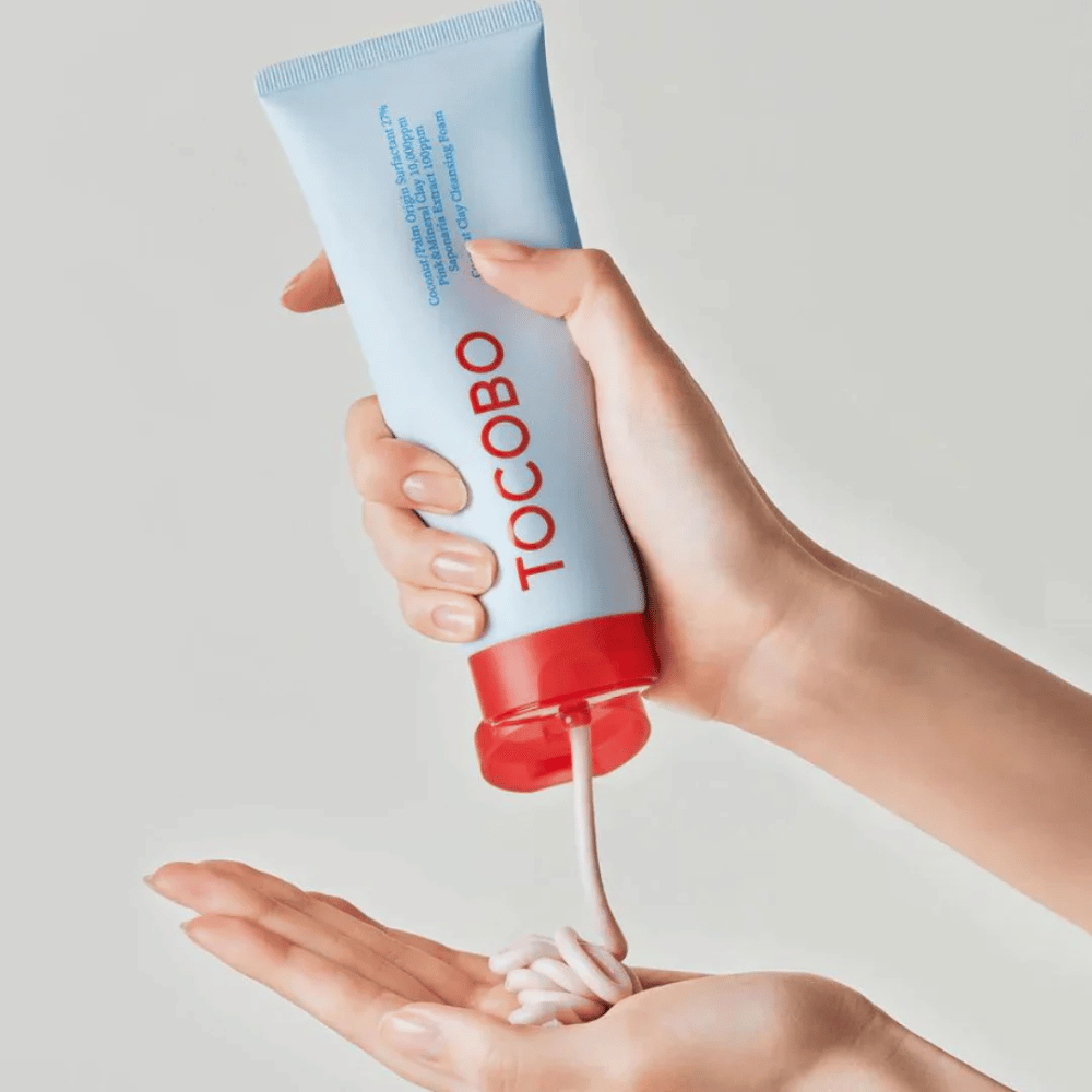 Una persona sostiene un tubo de TOCOBO Coconut Clay Cleansing Foam. El tubo es de color azul claro con una tapa roja y el texto "TOCOBO" en la parte delantera. El producto se exprime del tubo y se forma como un champú en la mano. El fondo es neutro y enfoca en el producto.