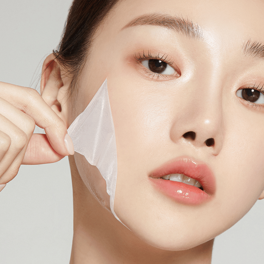 La imagen muestra a una joven quitándose una fina mascarilla facial, destacando su piel hidratada y suave. La mascarilla sigue los contornos del rostro, dando la impresión de ser un producto de cuidado de la piel efectivo y delicado. Su expresión es relajada.