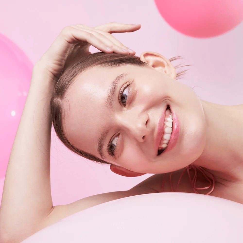 Una mujer sonríe y apoya su cabeza en la mano, rodeada de globos aerostáticos rosas. Su piel se ve radiante y naturalmente saludable.