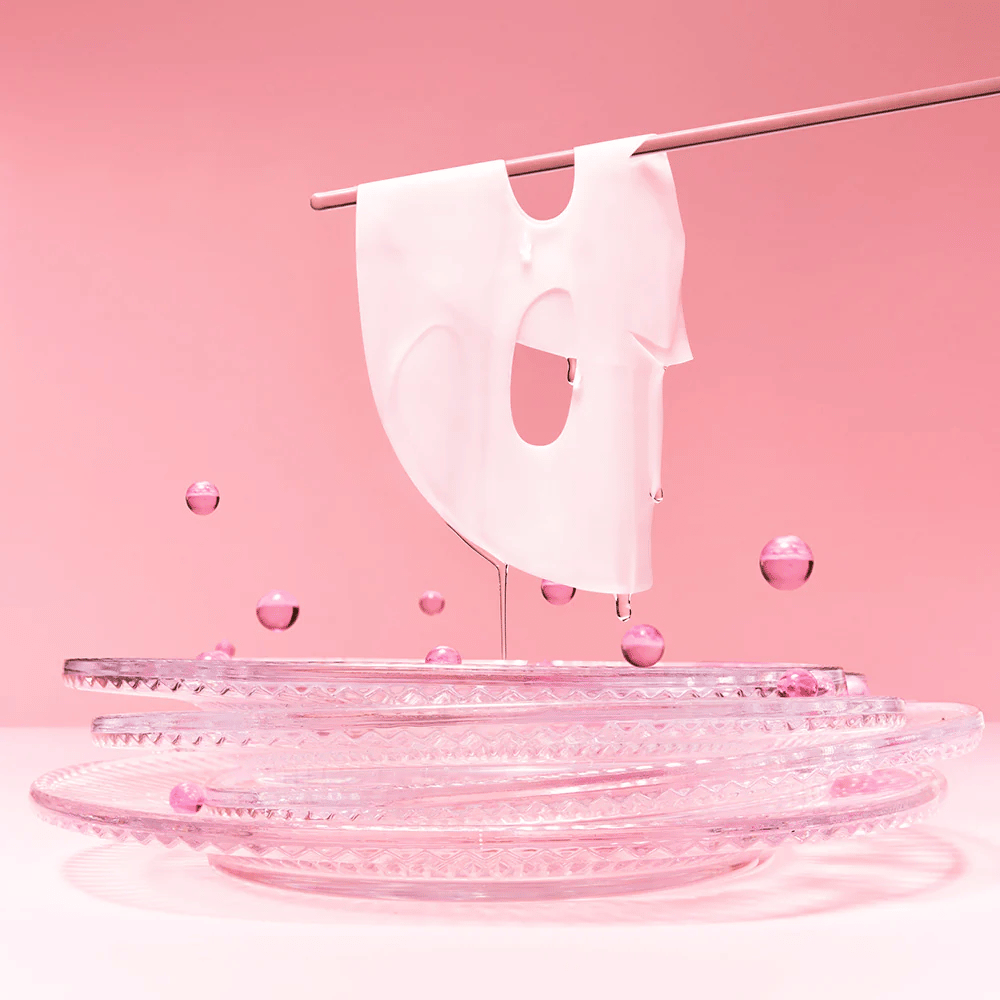 Una mascarilla de hidrogel cuelga de una barra, goteando con suero. El fondo es rosa y el espejo en la parte inferior refleja las burbujas rosas que rodean la mascarilla.