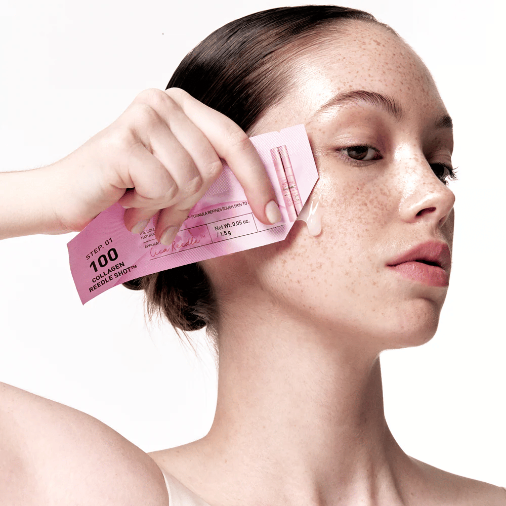 Mujer aplica el paso 1 de Collagen Reedle Shot 100 con un empaque rosa desechable en la mejilla. Un gel claro se ve en la piel, lo que marca el primer paso en el tratamiento de cuidado de la piel de dos pasos.