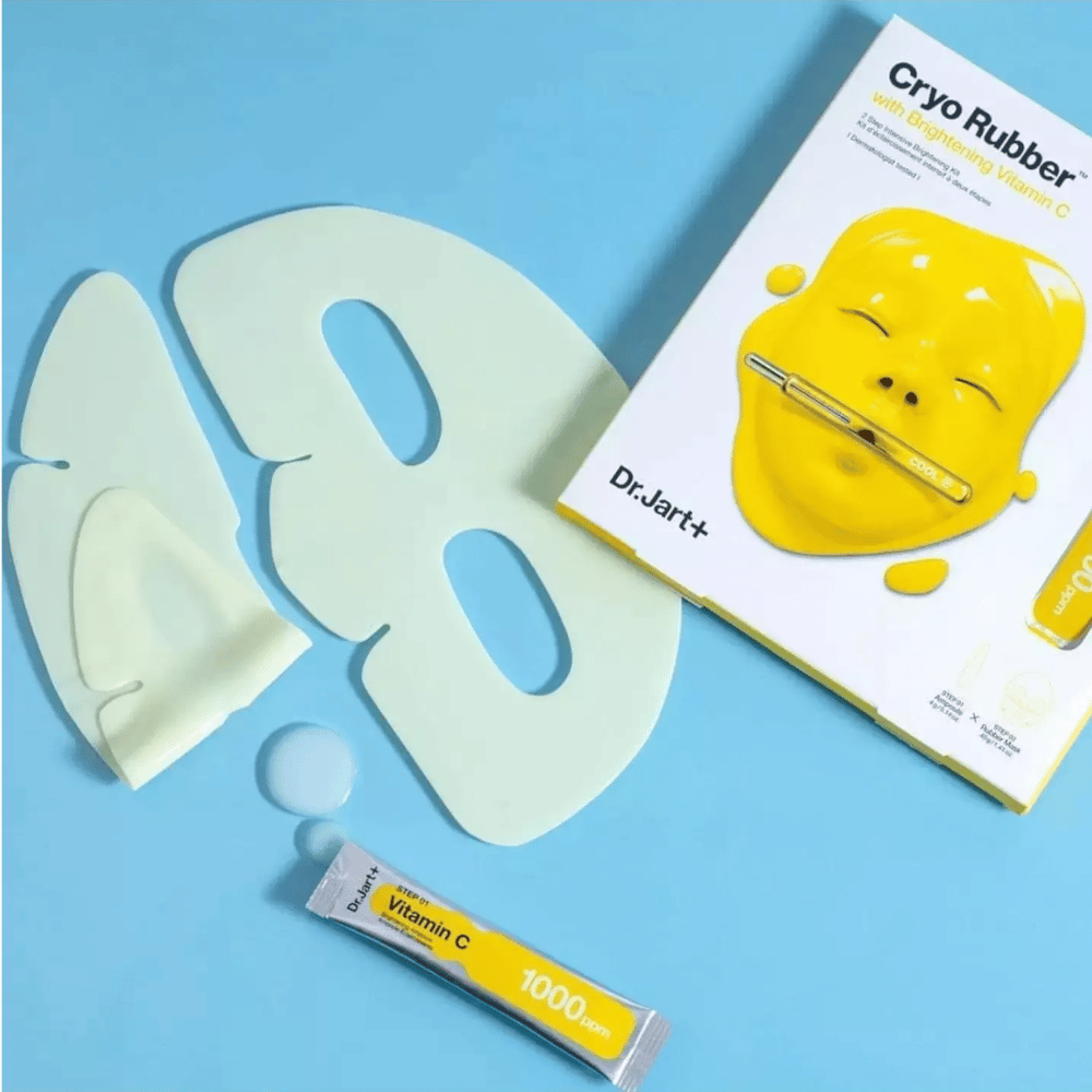 Imagen de Dr.Jart+'s 'Cryo Rubber™ con Vitamina C Iluminadora' producto de belleza. En un fondo azul se ve una mascarilla facial de goma blanca, un pequeño tubo marcado 'Vitamina C 1000ppm', y una caja con el nombre del producto y la imagen de la máscara amarilla.
