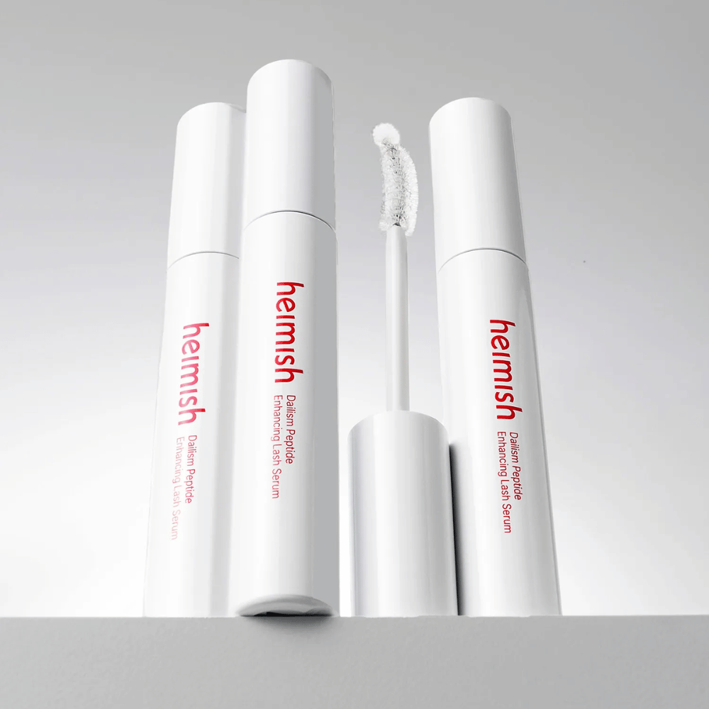 Se muestran varios envases de Heimish Dailism Peptide Enhancing Lash Serum junto con el cepillo aplicador curvado. La imagen destaca el elegante diseño del producto y el cepillo de precisión para una aplicación sencilla.