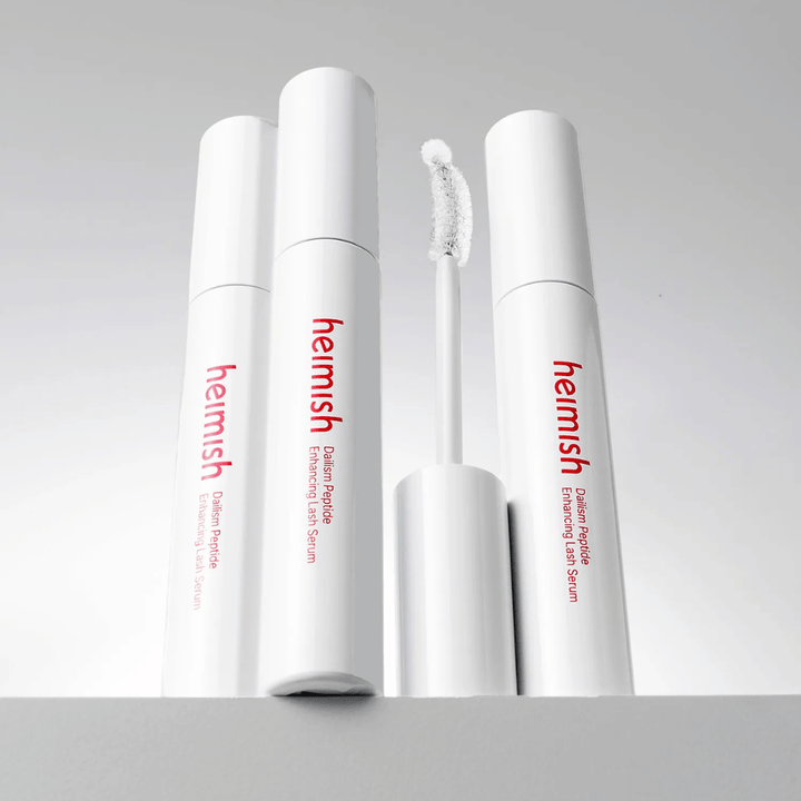 Se muestran varios envases de Heimish Dailism Peptide Enhancing Lash Serum junto con el cepillo aplicador curvado. La imagen destaca el elegante diseño del producto y el cepillo de precisión para una aplicación sencilla.