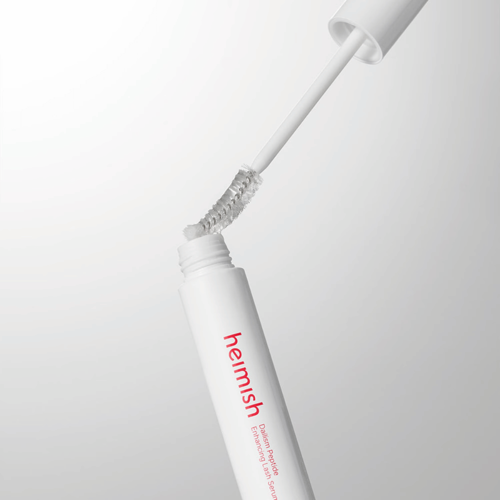 Primer plano del aplicador de Heimish Dailism Peptide Enhancing Lash Serum. El cepillo se extrae del recipiente blanco y muestra la fórmula transparente del sérum que se aplica sobre las pestañas.