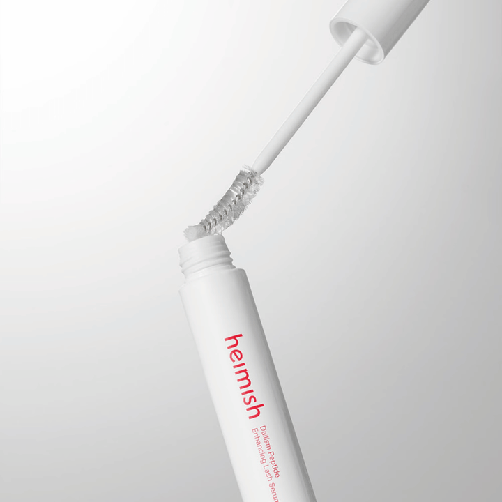 Primer plano del aplicador de Heimish Dailism Peptide Enhancing Lash Serum. El cepillo se extrae del recipiente blanco y muestra la fórmula transparente del sérum que se aplica sobre las pestañas.