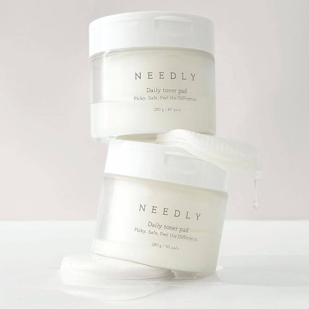 Dos frascos con Needly Daily Toner Pad apilados uno sobre otro. Los frascos son blancos con texto minimalista y contienen 60 almohadillas de tónico cada uno. Una almohadilla de tónico está parcialmente fuera del frasco superior con líquido goteando.
