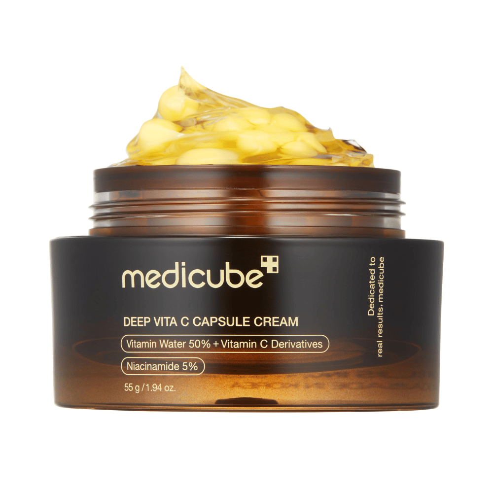 Un primer plano de "Medicube Deep Vita C Capsule Cream", que muestra la consistencia de la crema y las cápsulas de vitamina C. El empaque es de color marrón oscuro con texto dorado y describe el contenido de agua vitaminada (50 %) y niacinamida (5 %), lo que proporciona una presentación visual de las propiedades hidratantes y mejoradoras de la piel del producto.