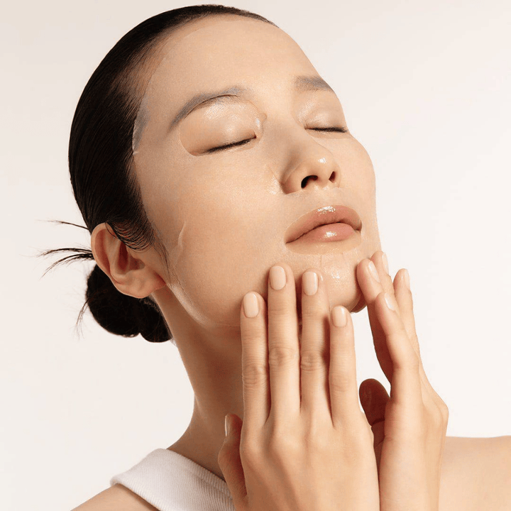 Modelo con piel hidratada y radiante aplica Sheet mask, enfoque en brillo, tono de piel uniforme y resplandor.