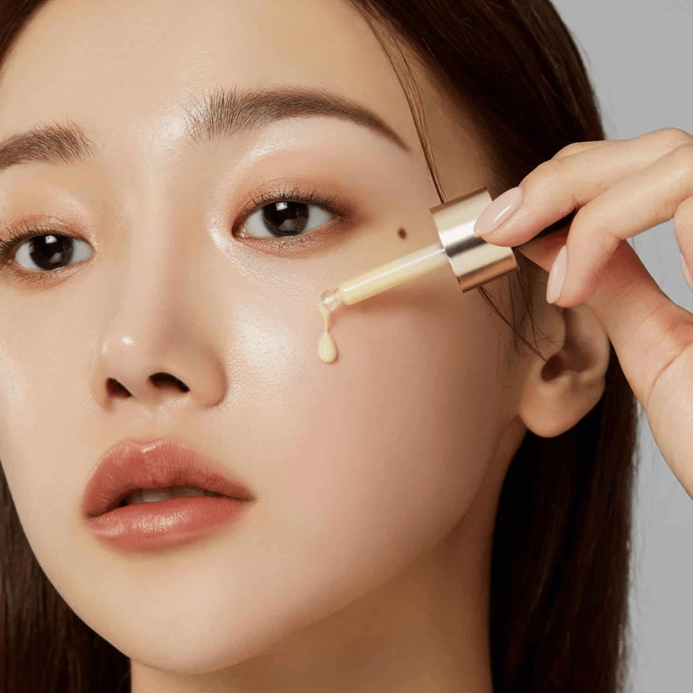 Primer plano de una mujer aplicando Medicube Deep Vita C Ampoule con una pipeta dorada. Una gota del suero está a punto de alcanzar su piel, que tiene un brillo natural. Perfecto para proporcionar un tono de piel más claro y uniforme.