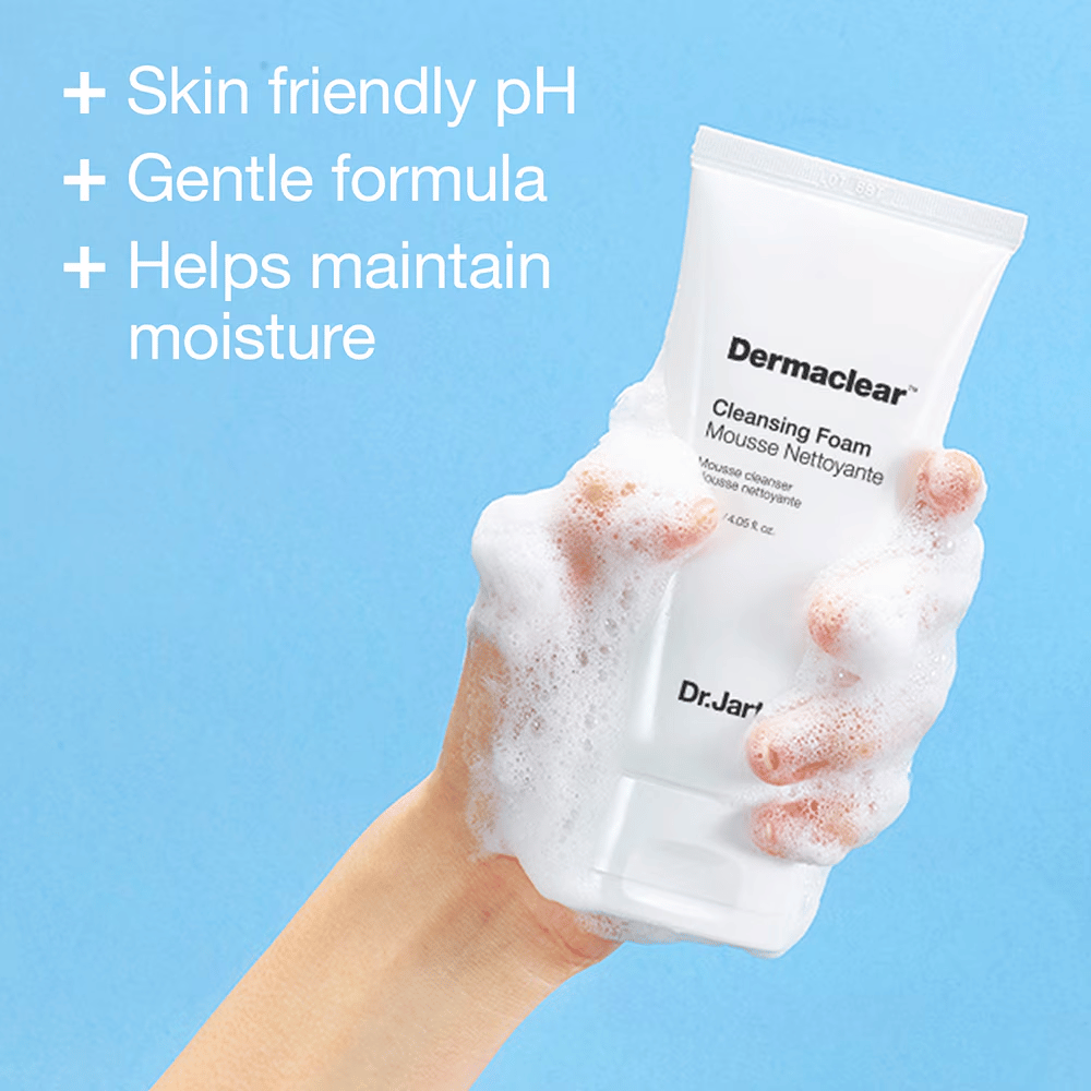 Mano cubierta de espuma sosteniendo Dr.Jart+ Dermaclear Cleansing Foam contra un fondo azul, con texto que destaca el pH amigable con la piel, fórmula suave y propiedades hidratantes.