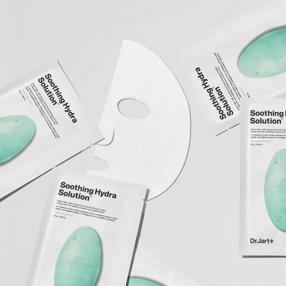 Varios paquetes de mascarillas "Soothing Hydra Solution" de Dr.Jart+ están esparcidos sobre una superficie clara. Una mascarilla desplegada está colocada en el centro, mostrando su estructura blanca y perforada.