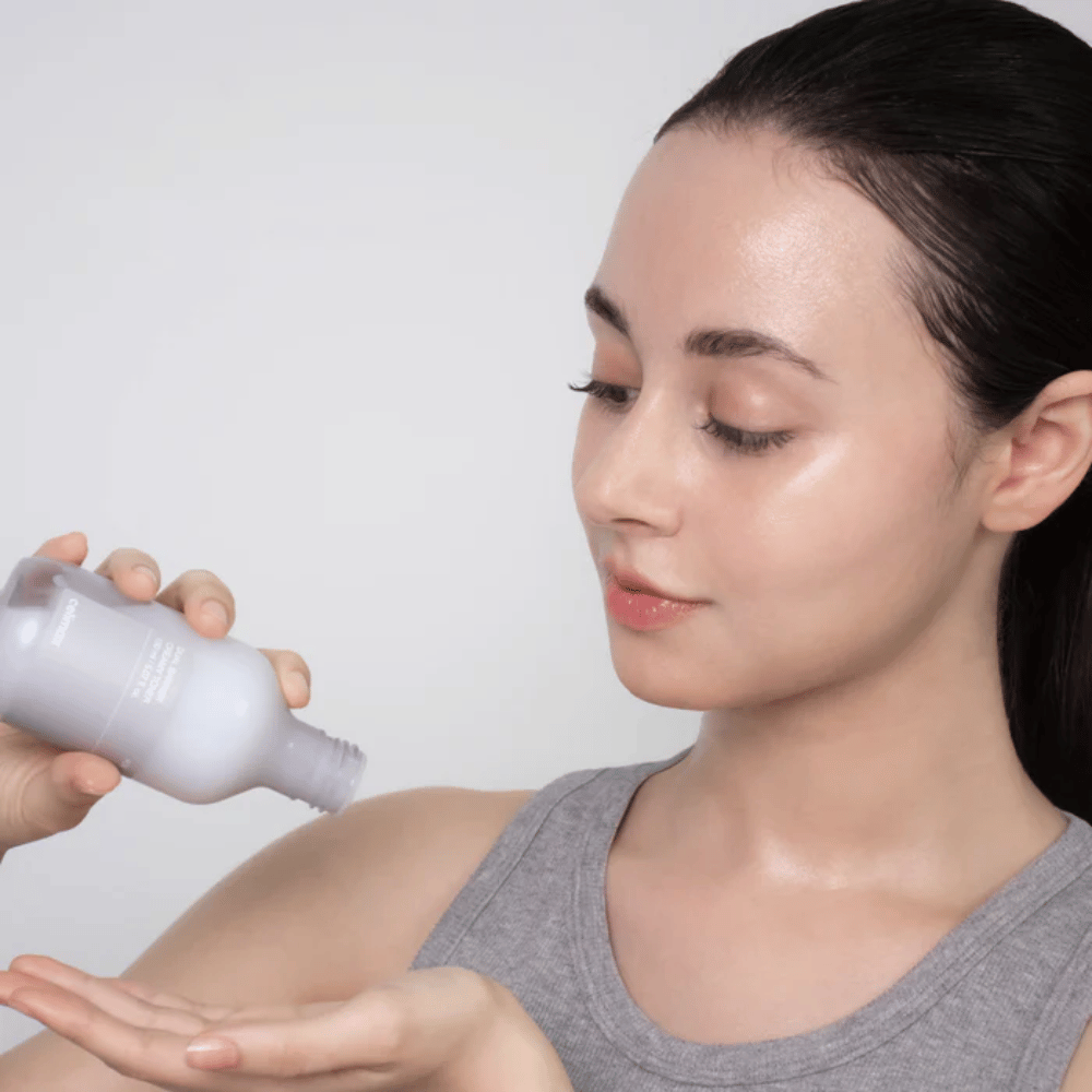 Mujer con piel brillante e hidratada aplica Celimax Dual Barrier Creamy Toner vertiendo el producto en la palma de la mano antes de su rutina de cuidado de la piel.
