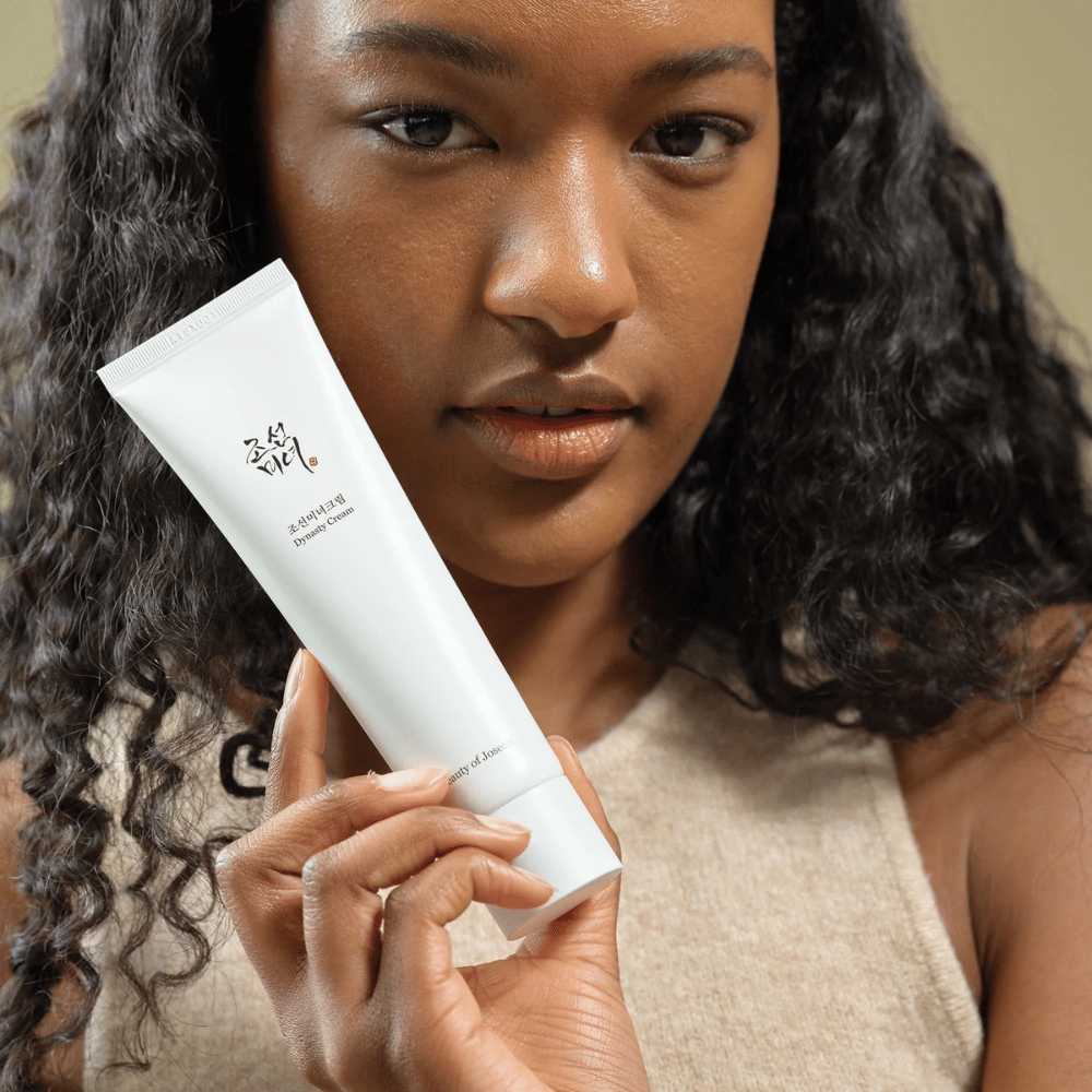 Una mujer con cabello rizado sostiene un tubo "Beauty of Joseon Dynasty Cream" cerca de su rostro, destacando el producto. La crema, presentada en un elegante tubo blanco, está diseñada para nutrir y rejuvenecer la piel, subrayando su atractivo para diferentes tipos de piel.