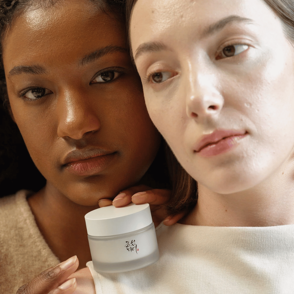 Dos mujeres, una con piel más oscura y otra con piel más clara, están de pie cerca una de la otra con enfoque en sus rostros y una crema facial de "Beauty of Joseon". La crema está colocada frente a ellas, destacando su uso para diferentes tipos de piel y su capacidad para dar brillo y nutrición.