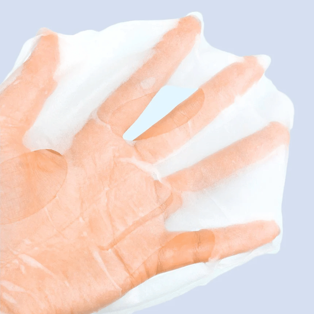Una mascarilla facial transparente y húmeda se sostiene en una mano contra un fondo azul claro. La máscara está diseñada para cubrir el rostro con espacios para los ojos y la boca.