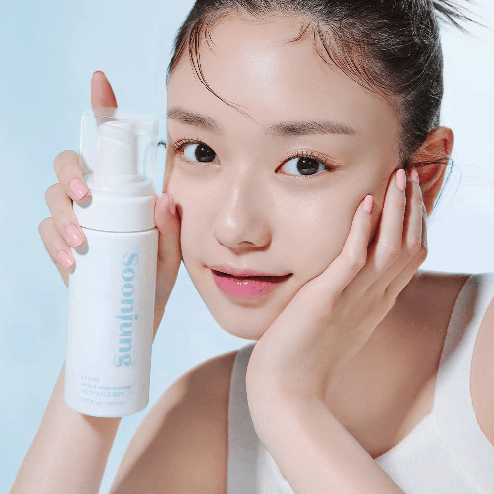 Una joven con piel clara sostiene una botella de bomba blanca con el texto "Soon Jung pH 6.5 Whip Cleanser" de ETUDE. Ella sonríe a la cámara con un maquillaje ligero y una apariencia fresca contra un fondo azul claro.