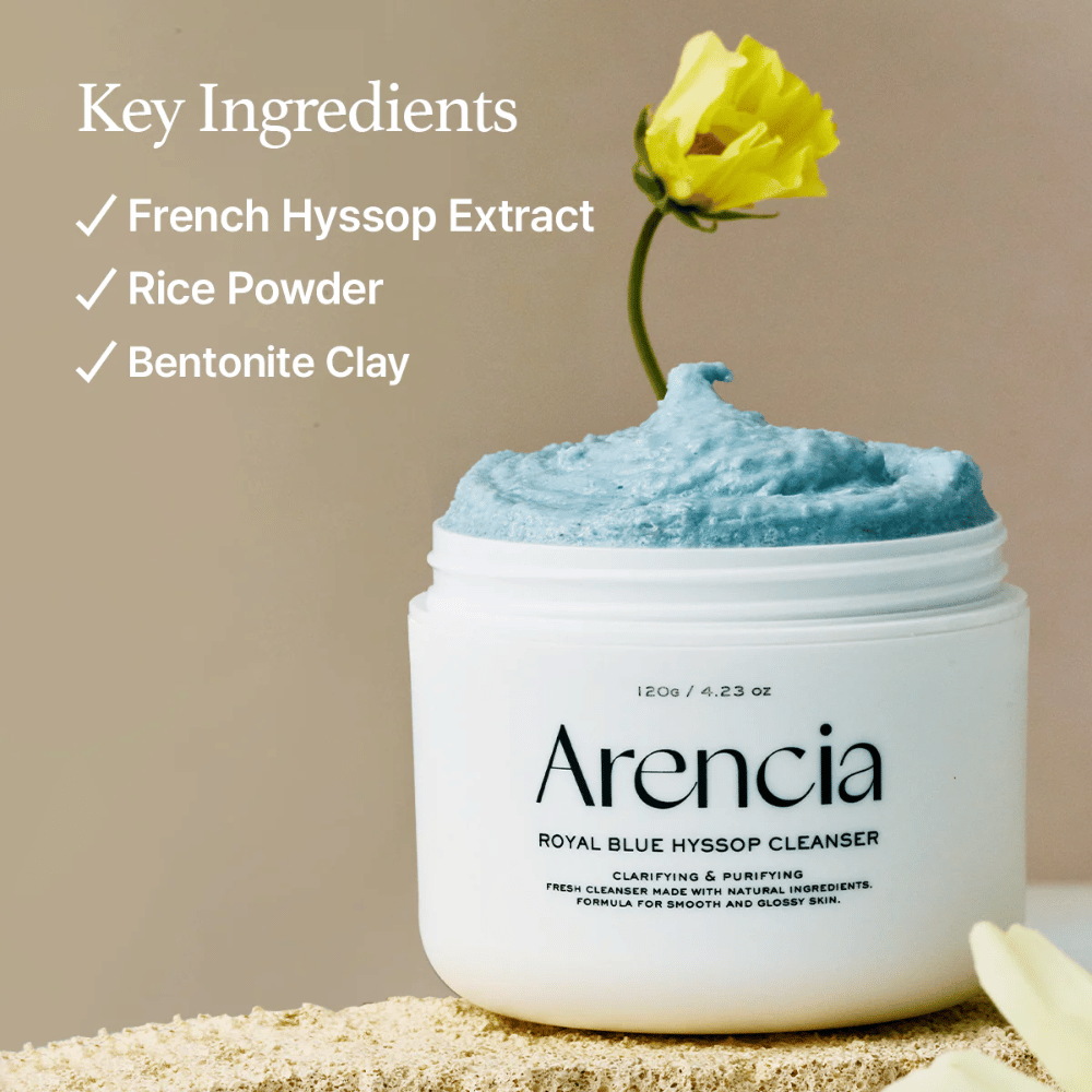 Imagen del frasco "Arencia Royal Blue Hyssop Cleanser" lleno de crema limpiadora azul. Una flor amarilla sobresale de la crema. El texto en la imagen destaca tres ingredientes clave: Extracto de Hisopo Francés, Polvo de Arroz y Arcilla de Bentonita.